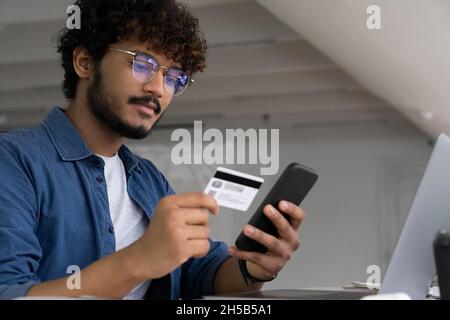 Young Indian man holding plastic bank card and smartphone make online payment Stock Photo