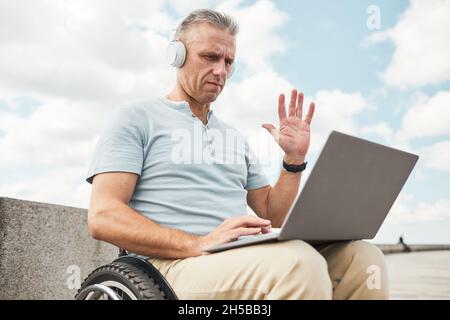 Portrait of adult man in wheelchair using laptop outdoors and waving at camera, copy space Stock Photo
