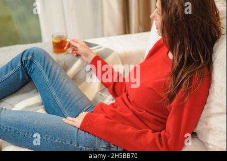Young woman relaxing on sofa holding cup with lemon tea. Woman dreaming in living room lying on couch. Woman thinking while drinking lemon tea at home Stock Photo