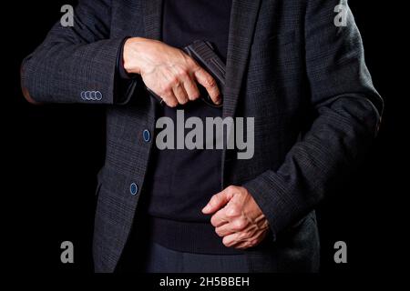 A man in a suit takes out a pistol from a jacket in darkness. Stock Photo