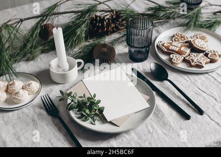 Festive Christmas table setting. Black cutlery, pine tree branch decoration. Gingerbread cookies, candleholder and glass. Linen tablecloth background Stock Photo