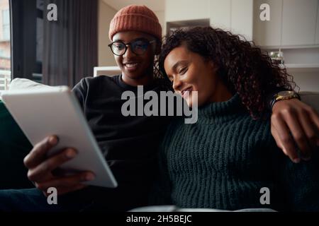 Smiling adult couple, multi-ethnic, sitting close together on sofa in modern apartment using tablet Stock Photo