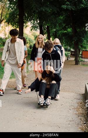 Teenage boy pushing cheerful girl sitting on skateboard while spending leisure time with friends in park Stock Photo