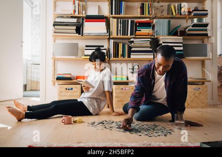 Man playing jigsaw puzzle while girlfriend reading book in living room Stock Photo