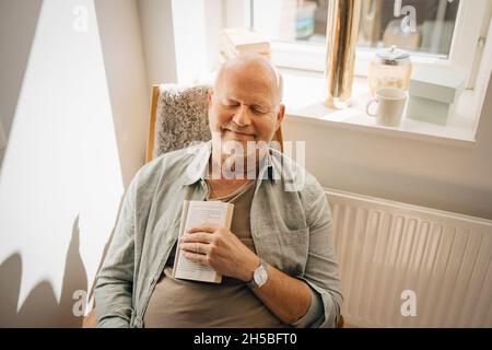 Smiling senior man with book relaxing on chair at home Stock Photo