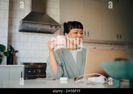 Smiling woman having coffee while on video call through laptop in kitchen Stock Photo