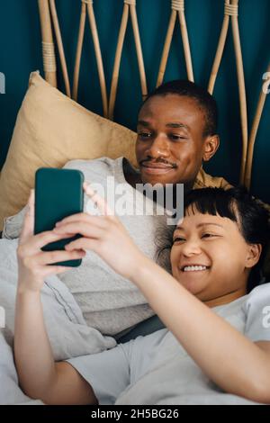 Smiling multiracial couple using mobile phone while lying on bed at home Stock Photo