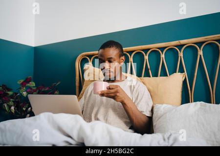 Mid adult man working on laptop while having coffee in bed Stock Photo