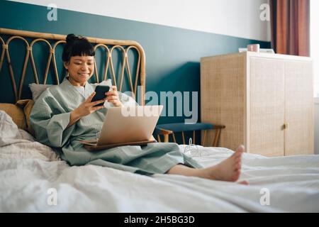 Smiling woman with laptop using smart phone on bed at home Stock Photo