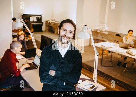 Portrait of smiling male programmer standing with arms crossed while colleagues working at desk in office Stock Photo