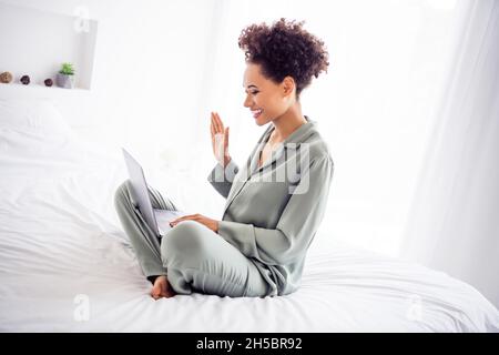 Profile side view portrait of attractive cheerful girl sitting on bed using laptop calling video at light white home indoors Stock Photo