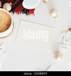 Christmas card design. Blank paper card mockup, cup of coffee, beige balls, scarf, bell on white table. Flat lay, top view, copy space Stock Photo