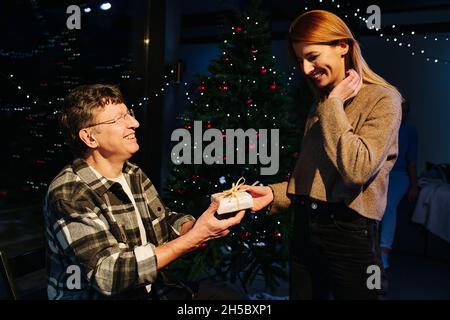 Lovely elder father and his adult daughter exchange christmas present. They are lit with a lamp, over dark decorated Christmas tree. Stock Photo