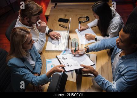 Four diverse financial analysts examining company graphs and charts during meeting at office room. Modern technology for work on table. People and cooperation concept. Stock Photo
