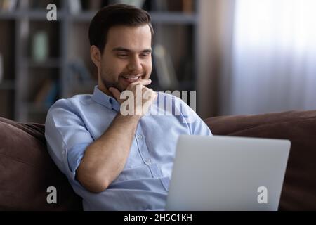 Smiling young man using computer, resting on sofa. Stock Photo