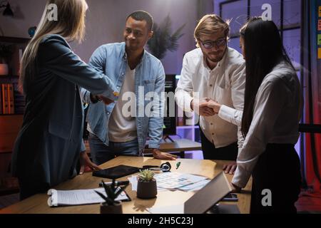 Team of multicultural economists shaking hands while standing near table with various gadgets and papers. Analysis of financial situation at company. Successful cooperation. Stock Photo