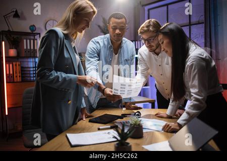 Business team of four brokers discussing financial statistic during evening conference at office. Multiracial partners standing around table and using modern technologies. Stock Photo