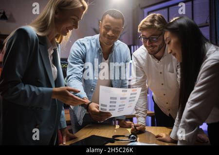 Business team of four experts discussing financial statistic during evening conference at office. Multiracial partners standing around table and using modern technologies. Stock Photo