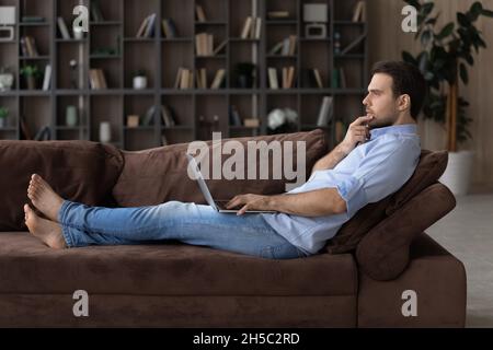 Pensive young man using computer, lying on sofa. Stock Photo