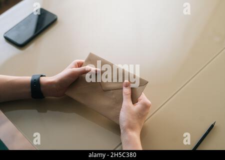 Close-up top view of unrecognizable young woman holding in hands envelope with received letter sitting at table. Stock Photo