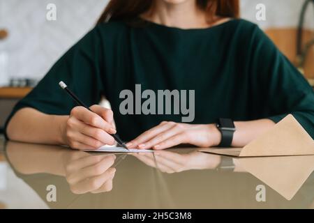 Close-up cropped shot of unrecognizable young woman writing handwritten letter sitting at table at home, selective focus, blurred background. Stock Photo