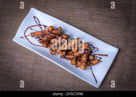 Presentation in a rectangular plate of aubergines cut into strips and fried, with sugar cane honey scattered on top, on a brown background. Stock Photo