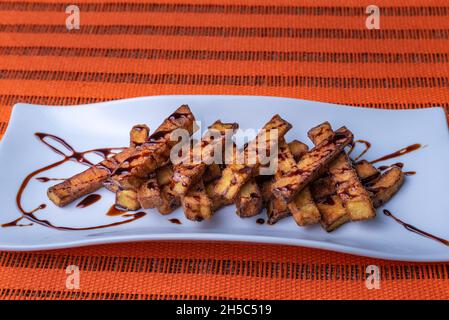 Presentation in rectangular plate of aubergines cut into strips and fried, with sugar cane honey scattered on top, on an orange background. Stock Photo