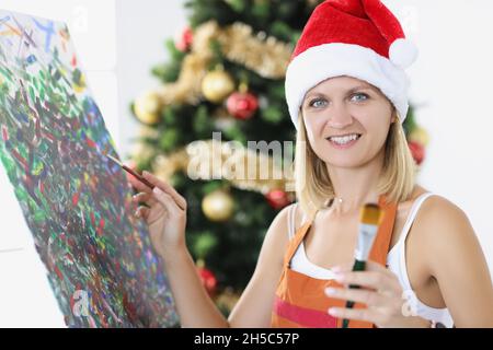 Portrait of young smiling woman artist painting picture against background of New Year tree