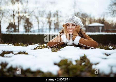 happy stylish middle aged woman with mittens in a knitted hat and sheepskin coat looking into the distance outdoors in the city park in winter. Stock Photo