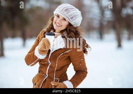 happy stylish 40 years old woman with mittens and cup of hot chocolate in a knitted hat and sheepskin coat outside in the city park in winter. Stock Photo