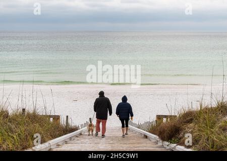 Seaside, USA - January 10, 2021: Couple people with dog walking in Seaside, Florida gulf coast beach with wooden boardwalk steps down and white sand w Stock Photo