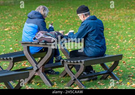 elderly couple seated at a wooden bench enjoying the fresh-air and a picnic with cup of tea, older couple taking a break and drinking tea out of doors. Stock Photo