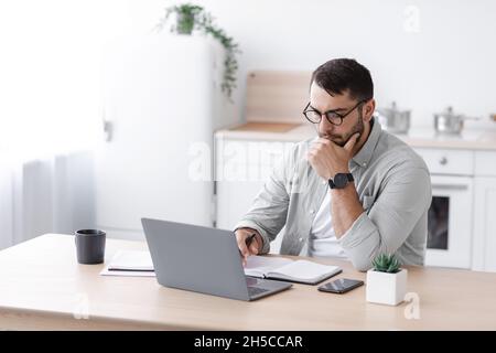 Busy pensive middle aged european businessman in glasses working at computer on minimalist kitchen interior Stock Photo