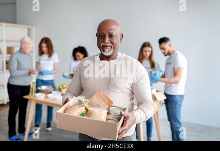 African american senior man holding box with donations food, looking and smiling at camera in charity center Stock Photo