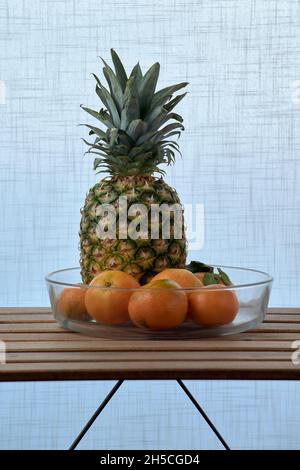 glass dish with pineapple and oranges, on a wooden slat table and a white textured background Stock Photo