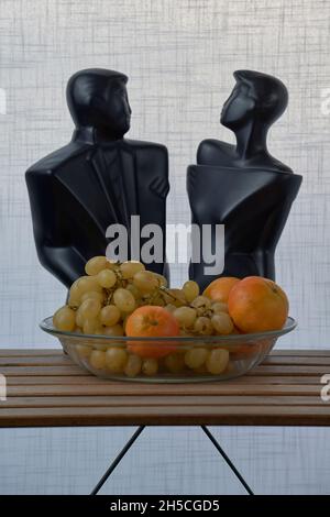 Glass plate with grapes, oranges and the black sculptures of a man and a woman, on a wooden slat table and a white textured background Stock Photo
