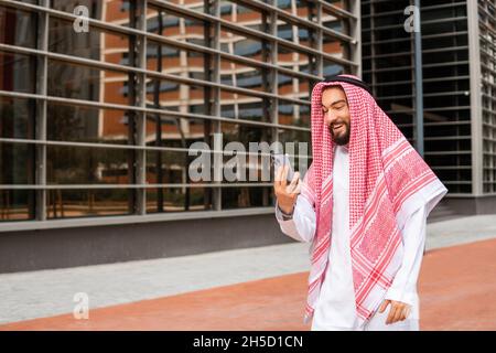 Happy Arab man in keffiyeh smiling and browsing smartphone while standing on city street Stock Photo