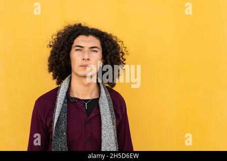 Short medium shot Portrait of a serious young Latino man in jean and burgundy shirt, with curly hair, standing in front of a yellow wall in the backgr Stock Photo