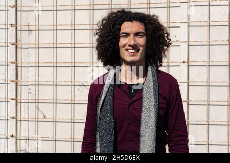 Short medium shot Portrait of a smiling young Latino in jean and burgundy shirt, with curly hair, standing in front of a wall with a metal grill. copy Stock Photo