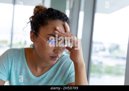Tired biracial businesswoman in eyeglasses sitting with head in hand at creative office Stock Photo