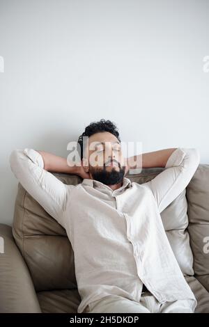 Tired man in white shirt closing eyes and leaning back on comfy sofa Stock Photo