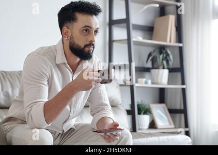 Serious handsome man drinking cup of espresso and watching news or show on tv at home Stock Photo