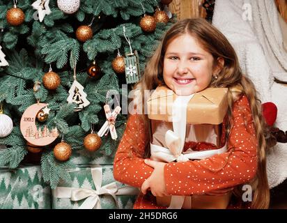 Young happy smiling long haired girl opening her Christmas gift on decorated Christmas tree background.. Girl at Xmas. Rustic concept. Stock Photo