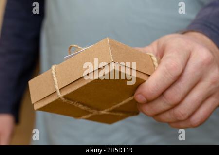 A man's hand holds out a brown gift box to the camera. The rectangular cardboard box is tied with string. Close-up. Selective focus. Stock Photo