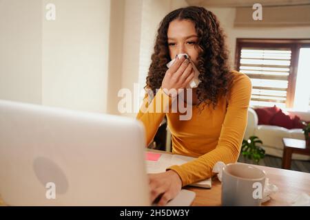 Multi-cultural female typing from home on laptop whilst blowing her nose. Sickness whilst working from home Stock Photo