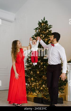 New year and Christmas. Mom and dad hold hands with their little son in the background of the Christmas tree and lift him up Stock Photo