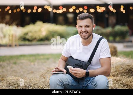 Happy young man sitting on hay bench and using wireless headphones. Man laughing and have fun Stock Photo
