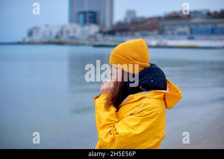 lonely woman in yellow raincoat freezes on sea shore against backdrop of winter cityscape in cold blue tones Stock Photo