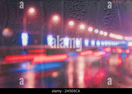 Blurry lights of moving cars and lanterns reflecting on the wet asphalt in the night city behind rain-covered window. Rainy bad autumn weather concept Stock Photo