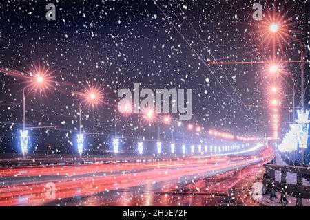 Traces of headlights from cars moving at winter night on the bridge, illuminated by lanterns in a snowfall. Lights reflecting in the wet asphalt. Stock Photo